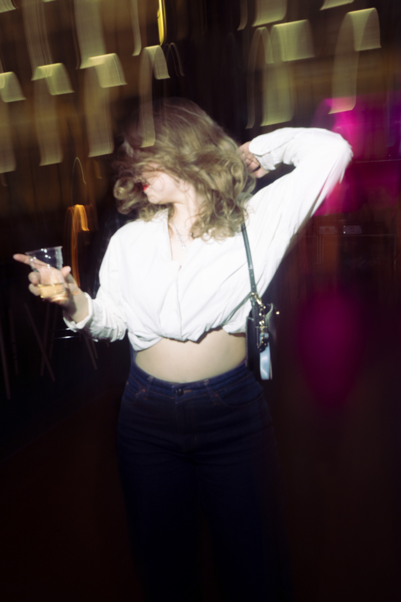 Person with shoulder-length hair dancing, wearing a white cropped shirt, dark jeans, holding a drink in one hand, blurred motion and colorful light streaks in the background.