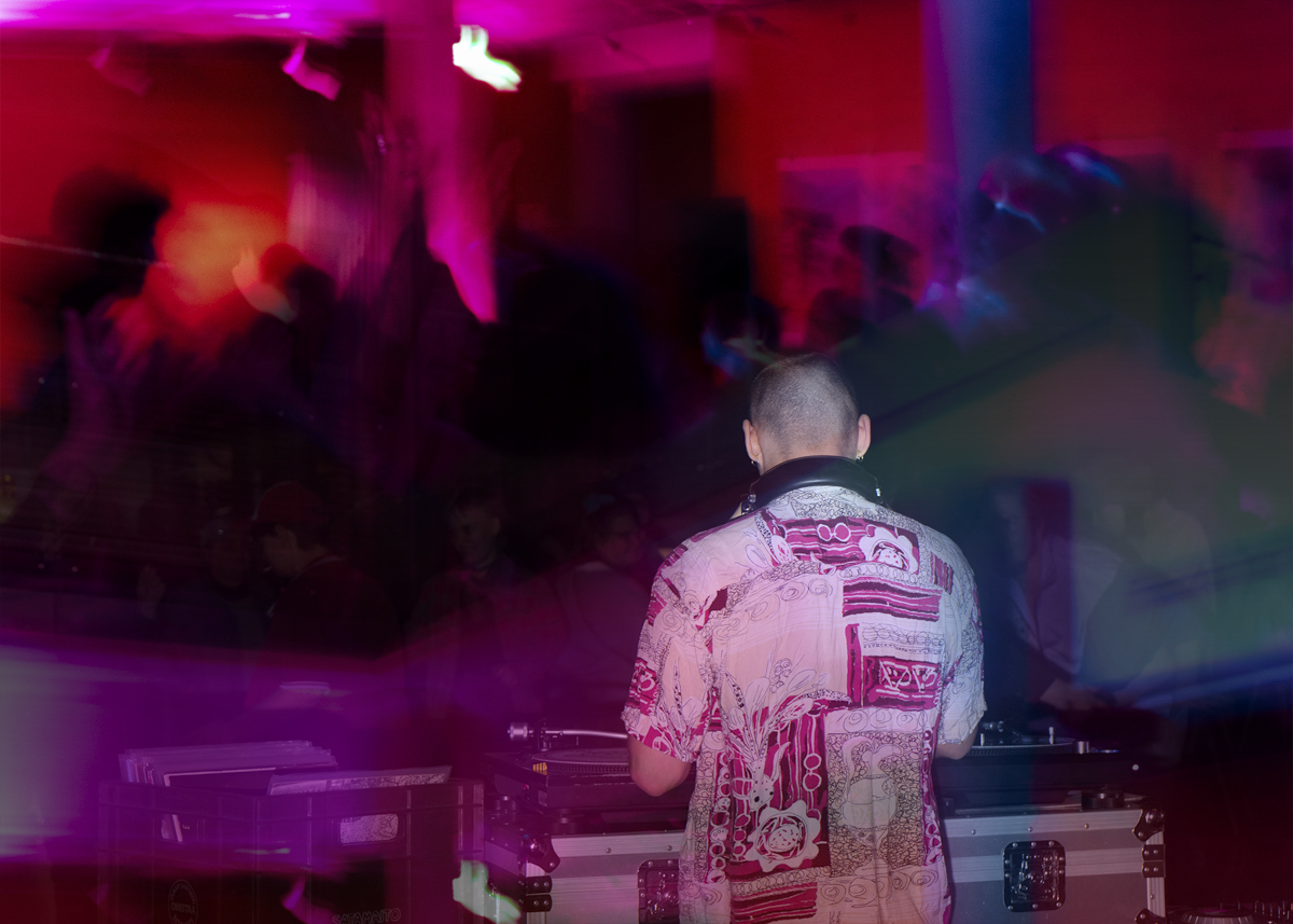 Rear view of a DJ wearing headphones and a patterned shirt playing music on DJ equipment in a dimly lit club with pink and purple lights and blurred crowd.