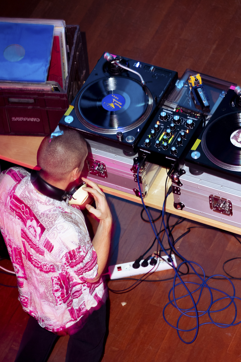 DJ wearing a pink and white patterned shirt using headphones with one hand on a turntable in front of him on a wooden floor, next to another turntable and a mixer with colorful knobs and buttons, with a crate of vinyl records nearby.