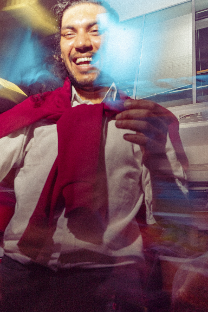 Smiling man with curly hair wearing a white shirt and a red sweater draped over his shoulders, captured with light trails and blurs creating a dynamic and colorful effect.