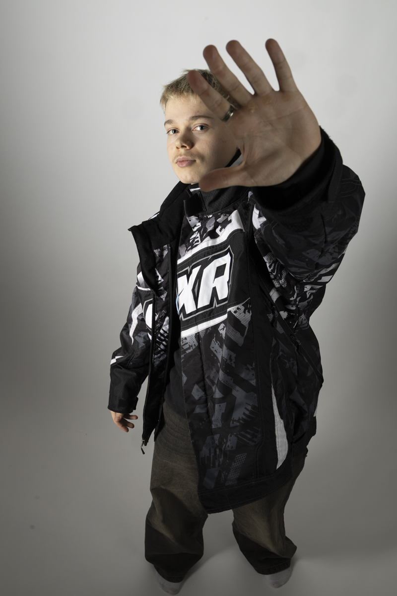 Person with short blonde hair wearing a black, white, and gray patterned jacket with 'FXR' on the front holds their hand up toward the camera, partially obscuring their face, against a plain light gray background.
