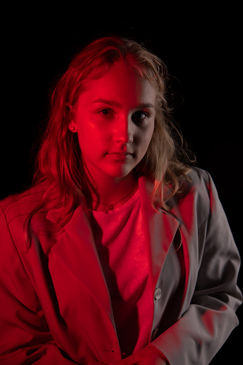 Portrait of a young woman with long blonde hair wearing a light-colored blazer and white shirt, illuminated by red lighting on a black background.