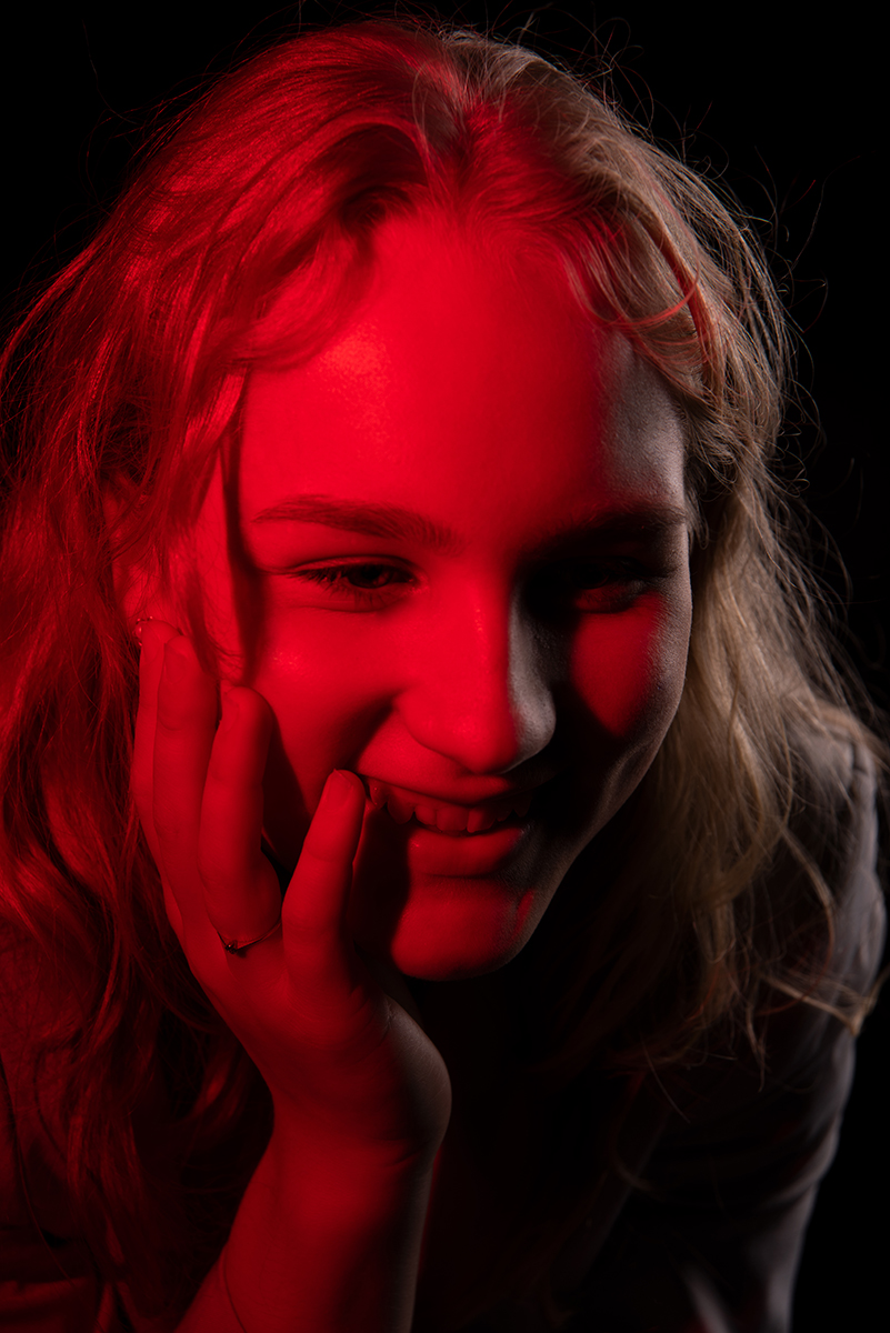 Close-up portrait of a young woman smiling with her hand gently touching her cheek, lit by dramatic red light against a black background.