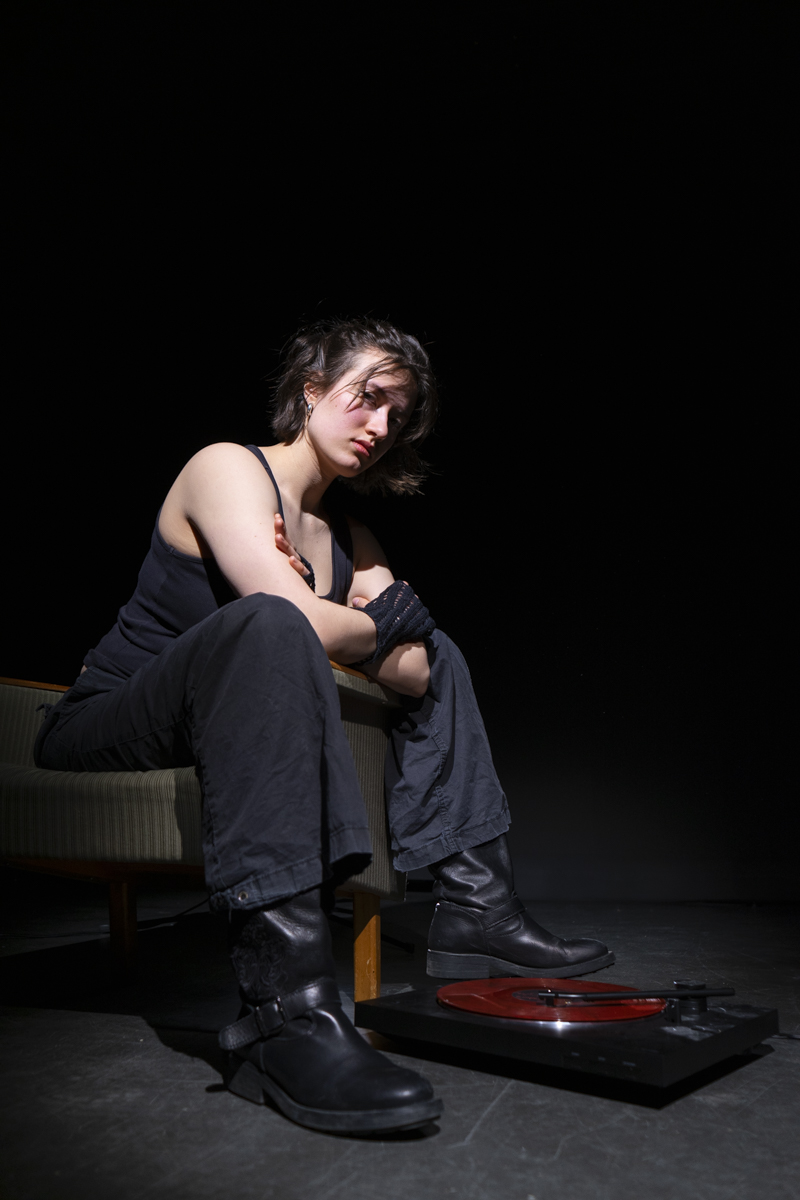 Young person with short hair wearing a black tank top, black pants, and black boots, sitting on a chair in a dark room with their arms crossed and looking downward, next to a red vinyl record on a black turntable on the floor.