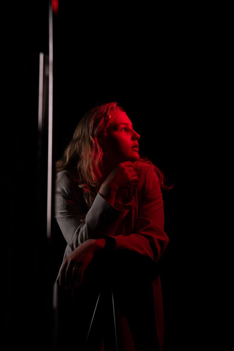 Woman with long hair lit by red light, gazing thoughtfully to her right, against a black background.