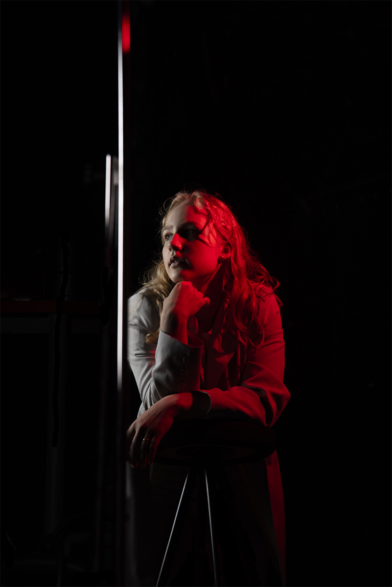 Young woman with long blonde hair leaning on a reflective vertical surface, partially illuminated by dramatic red light on her face and upper body against a dark background.