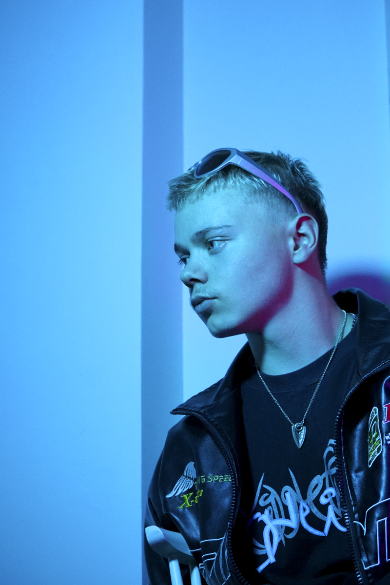 Young person with short hair wearing sunglasses on head, a dark jacket with patches, a graphic t-shirt, and a necklace, illuminated by blue lighting and looking to the left.