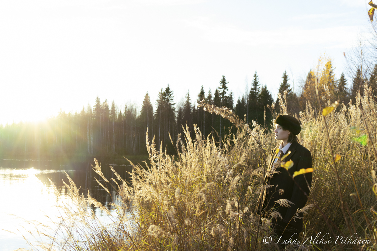 Person wearing a dark coat and hat standing among tall golden grasses by a reflective lake at sunset with a forest in the background.