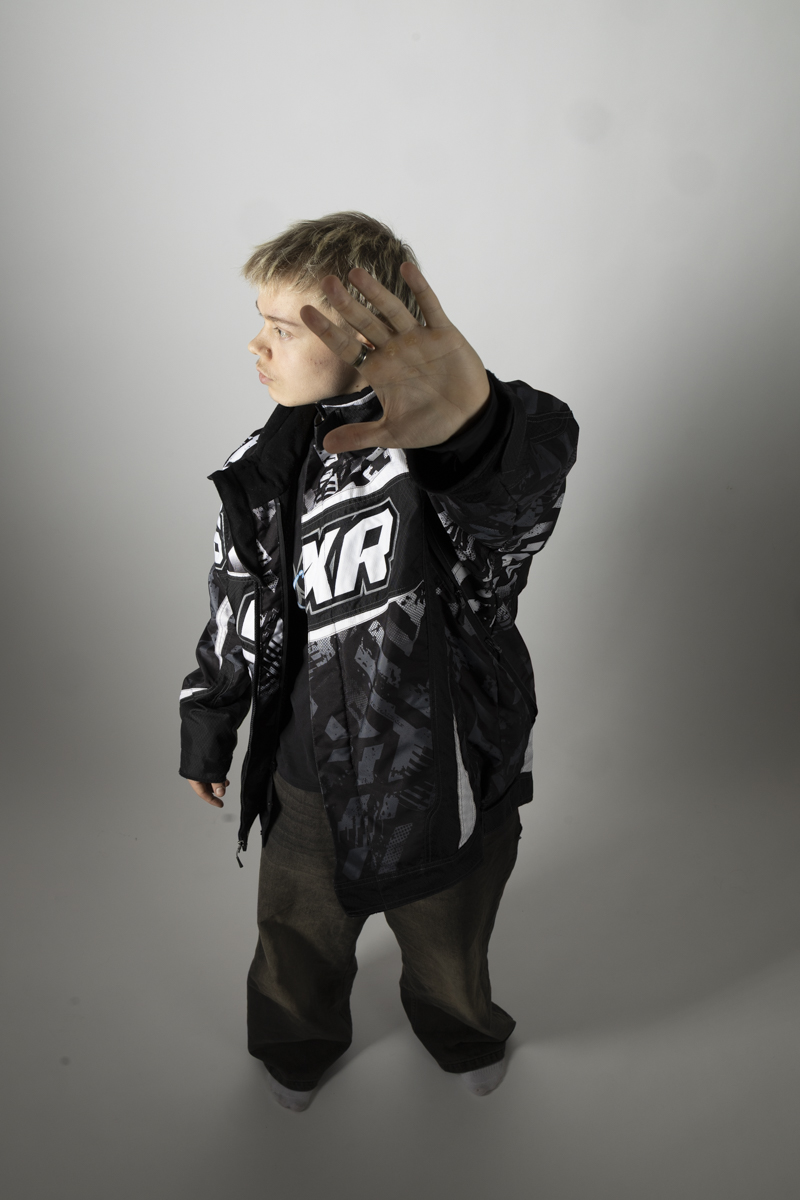 Person with short blonde hair wearing a black and white jacket with bold lettering, raising their left hand toward the camera as if to block the view, standing against a plain light gray background.