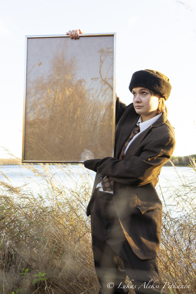 Person wearing a dark suit, patterned tie, and a black fur hat stands outdoors near a body of water, holding up a large framed mirror that reflects dry tall grass and trees.