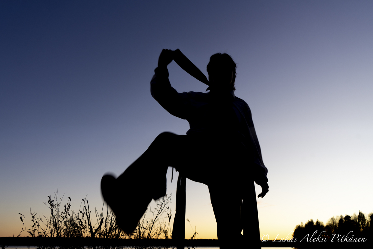 Silhouette of a person standing on one leg with the other leg raised, holding an object in the raised hand against a clear sunset sky with some plants and trees in the background.