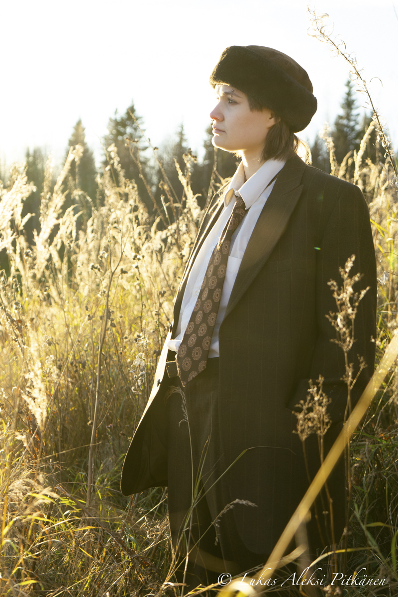 Person wearing a dark pinstripe suit, white shirt, patterned tie, and a black fur hat, standing in a sunlit field of tall dry grasses with conifer trees in the background, looking contemplatively to the right.
