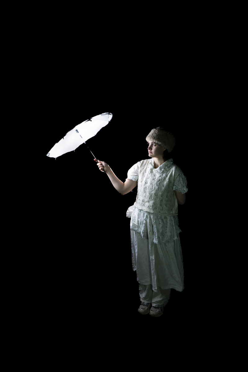 Person dressed in white vintage-style clothing and a furry hat holding a white umbrella against a black background, looking slightly to the side.