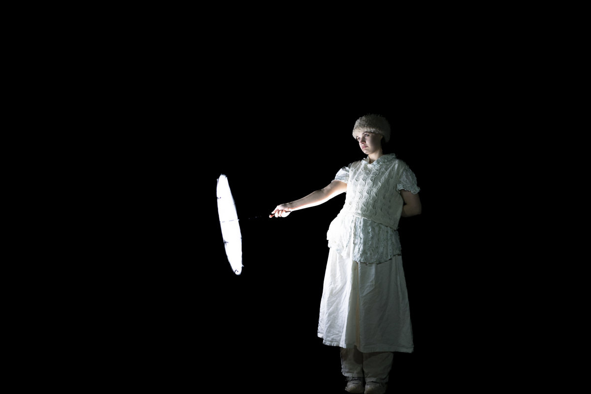 A person dressed in white clothing and a hat stands against a black background, holding a brightly lit white umbrella to their side.