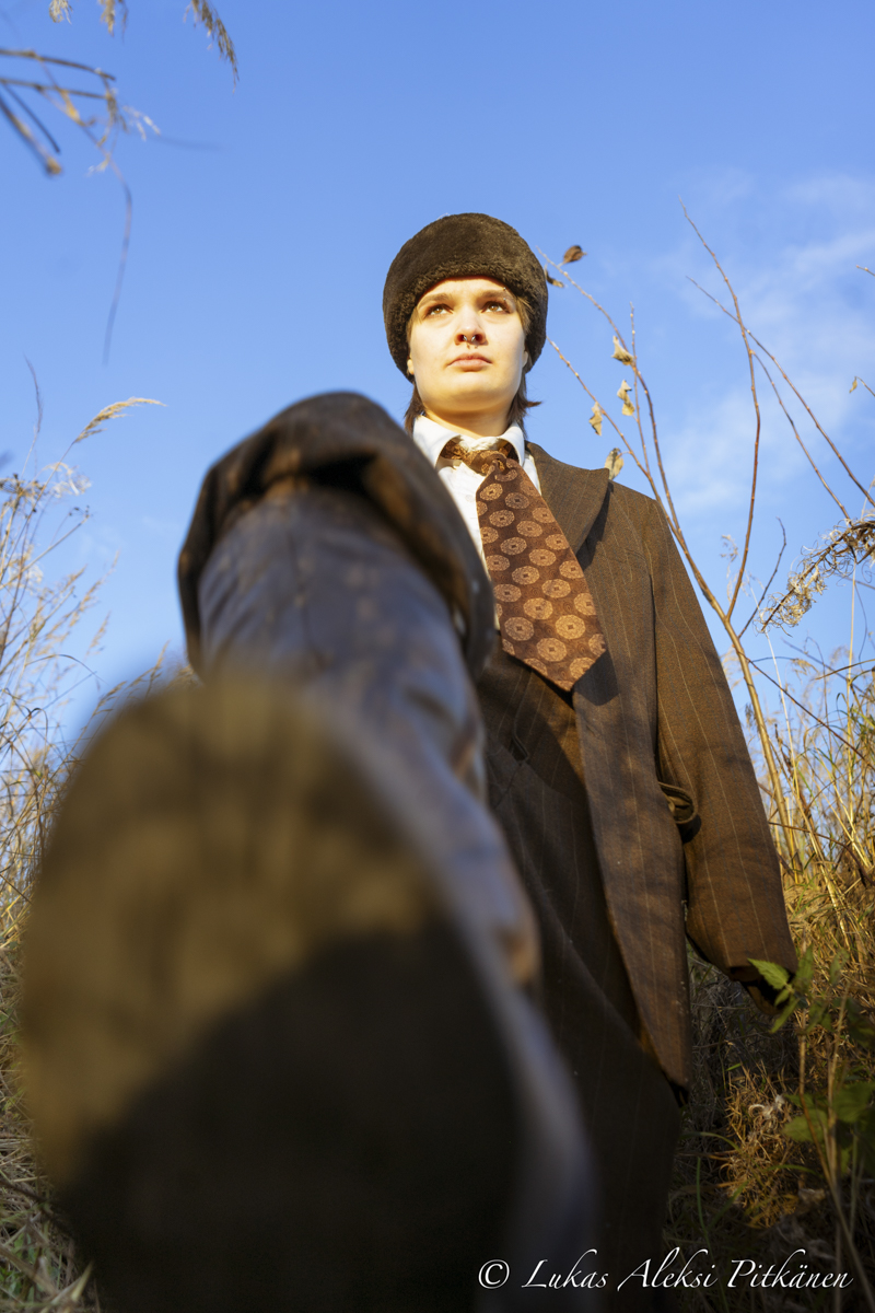 Person wearing a brown suit, patterned tie, and dark fur hat, taken from low angle with one foot extended toward the camera against a blue sky and dry plants.