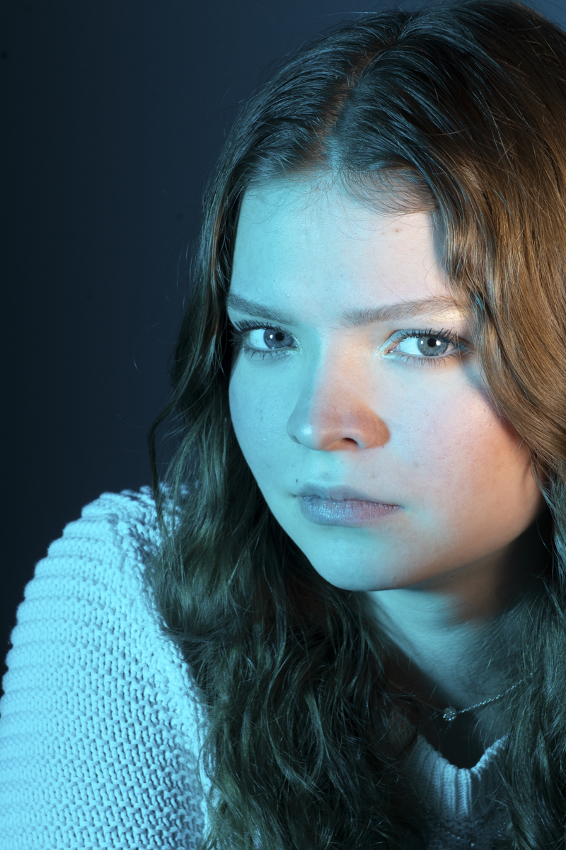 Close-up portrait of a young woman with long wavy hair, wearing a white knit sweater, illuminated by blue light on a dark background, looking directly at the camera with a serious expression.