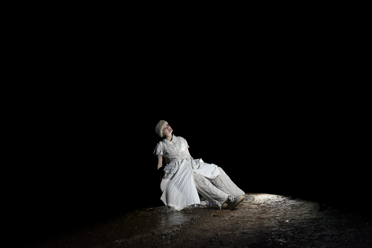 Person in white layered clothing and a textured hat sitting on the ground in a dark space, illuminated by a spotlight.
