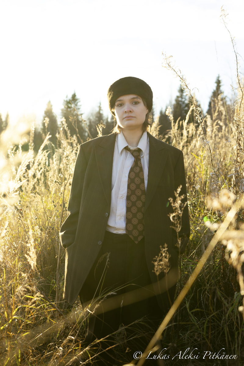 Person wearing a black fur hat, white shirt, patterned tie, and dark pinstripe suit jacket standing in a sunlit field of tall dry grass with trees in the background.