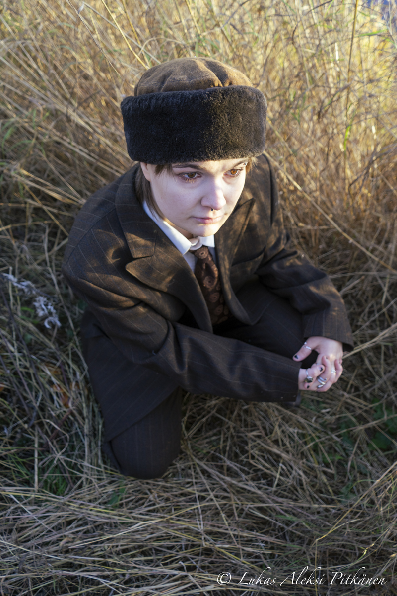 Person with short hair wearing a brown checkered suit, patterned tie, and a brown fur hat, sitting on dry grass surrounded by tall dry plants, looking slightly downward with a neutral expression.