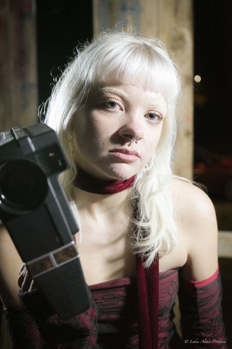 Portrait of a young person with platinum blonde hair and a septum nose ring, wearing a dark red scarf and off-shoulder top, holding a large black object close to the face, against a dark background with wooden beams.