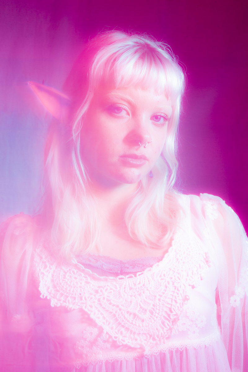 Portrait of a person with pale blonde hair, a septum piercing, and elf-like pointed ears, wearing a white lace top, bathed in pink and purple light.