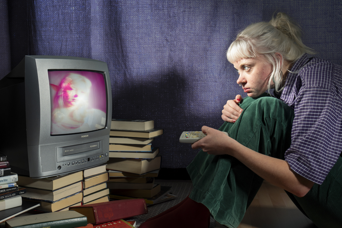 A young woman with platinum blonde hair, wearing a purple checkered shirt and green pants, sits on the floor with knees drawn up, intently watching an old CRT television with a remote in hand, surrounded by stacks of books.