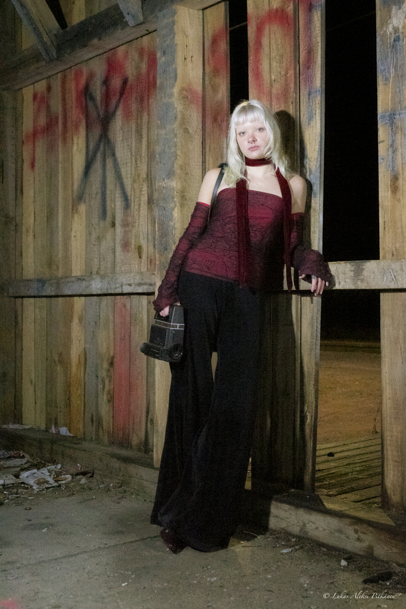 A woman with long blonde hair and bangs stands against a wooden wall with graffiti, holding a vintage camera in her right hand. She wears a burgundy strapless top, matching fingerless gloves and scarf, and loose black pants. The setting is dimly lit and appears rustic or abandoned with debris on the ground.