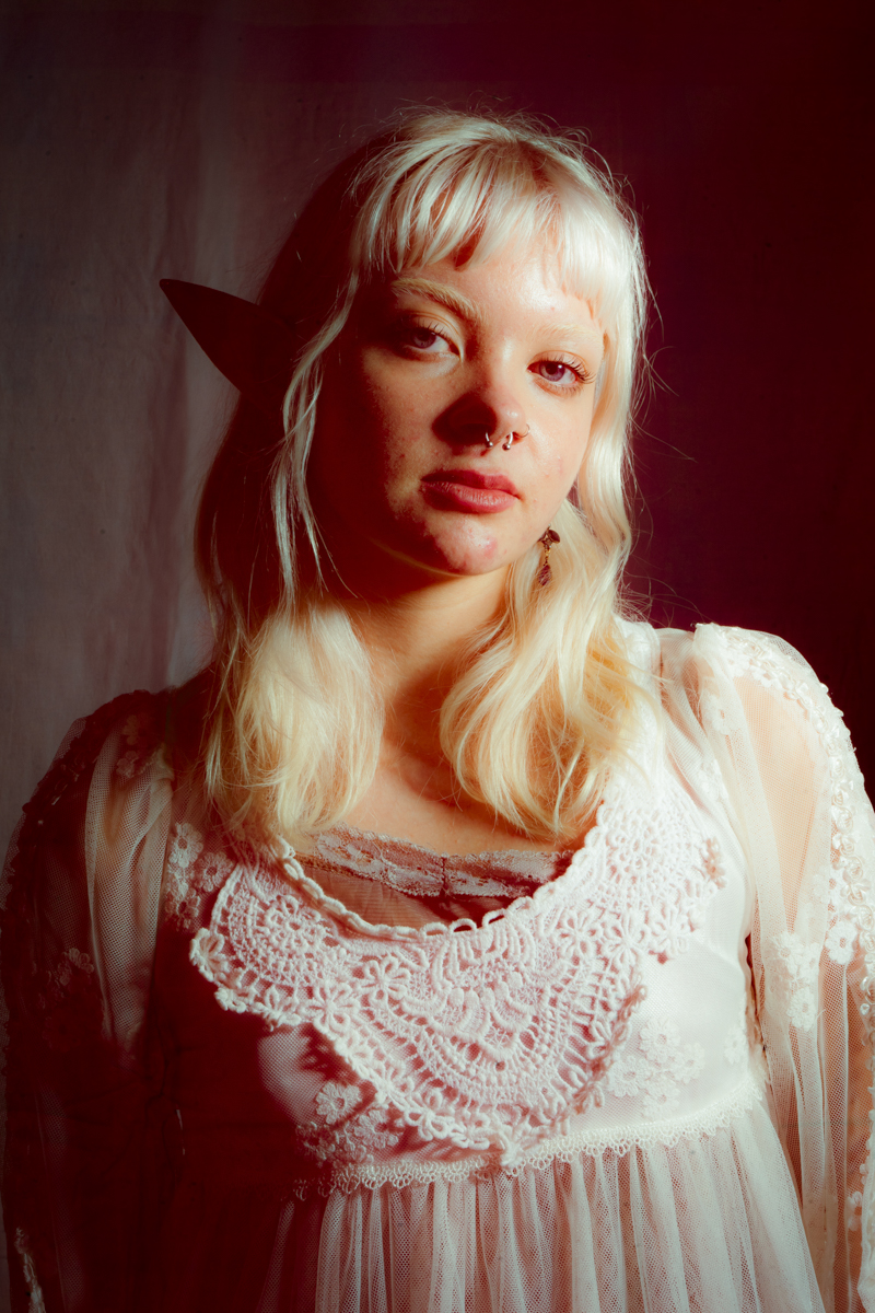 Young person with platinum blonde hair, wearing a white lace dress and a septum ring, posing against a dark background with elf-like pointed ear accessories.