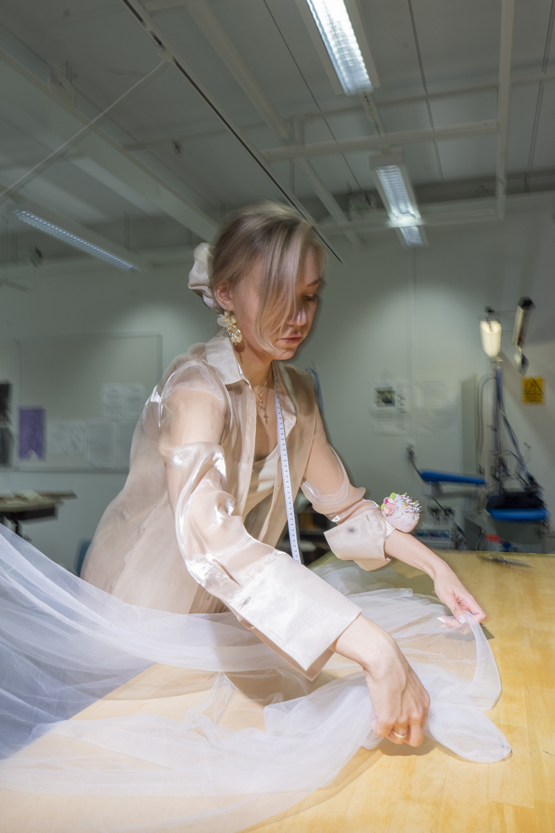 Young woman with a measuring tape around her neck and a pin cushion on her wrist arranging sheer white fabric on a wooden table in a brightly lit workshop.