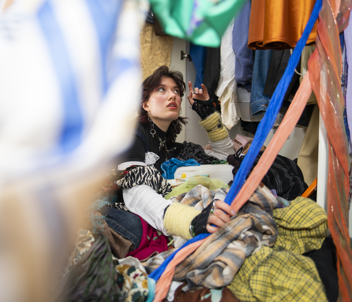 Young person with short dark hair and large earrings sitting among a large pile of mixed colorful clothes inside a closet, looking upward and holding some fabric with one hand.