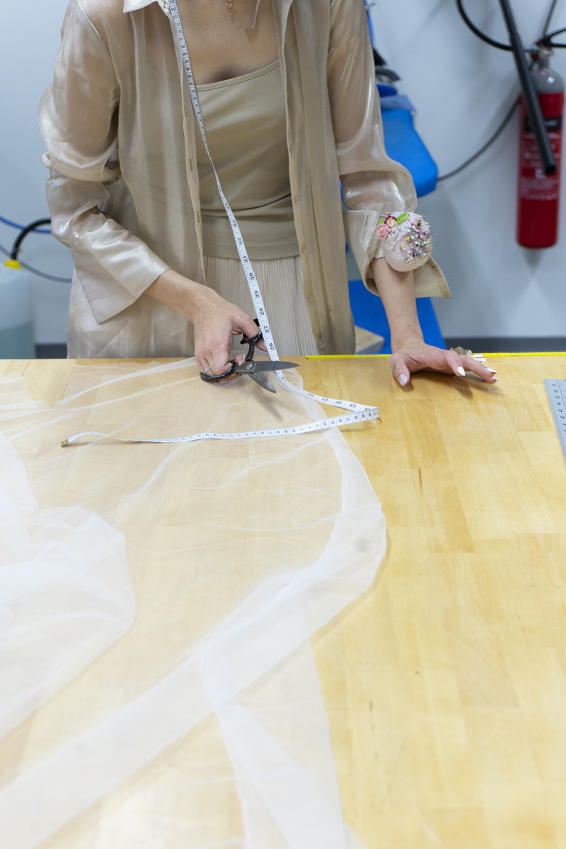 Person cutting sheer fabric on a wooden table with scissors while holding a measuring tape, wearing a sheer beige shirt with a pin cushion bracelet.