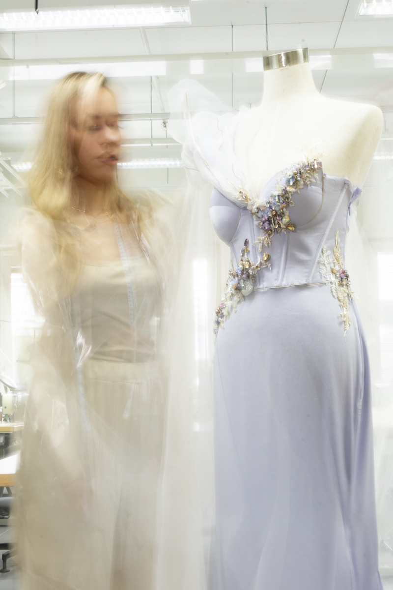 Blurry image of a woman with long blonde hair wearing a measuring tape around her neck standing next to a mannequin dressed in an elegant lavender dress decorated with floral and bead embellishments.