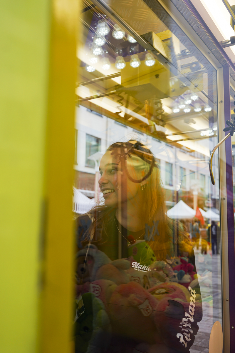 Young woman smiling inside a glass claw machine filled with colorful stuffed toys, reflected city buildings and outdoor tents in the background.