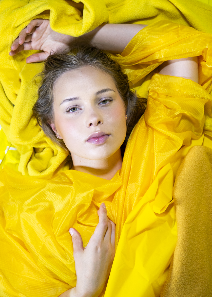 Close-up portrait of a young woman with light skin and green eyes lying on a bright yellow fabric background, wearing a yellow textured garment, her right arm bent above her head and left hand resting on her chest.