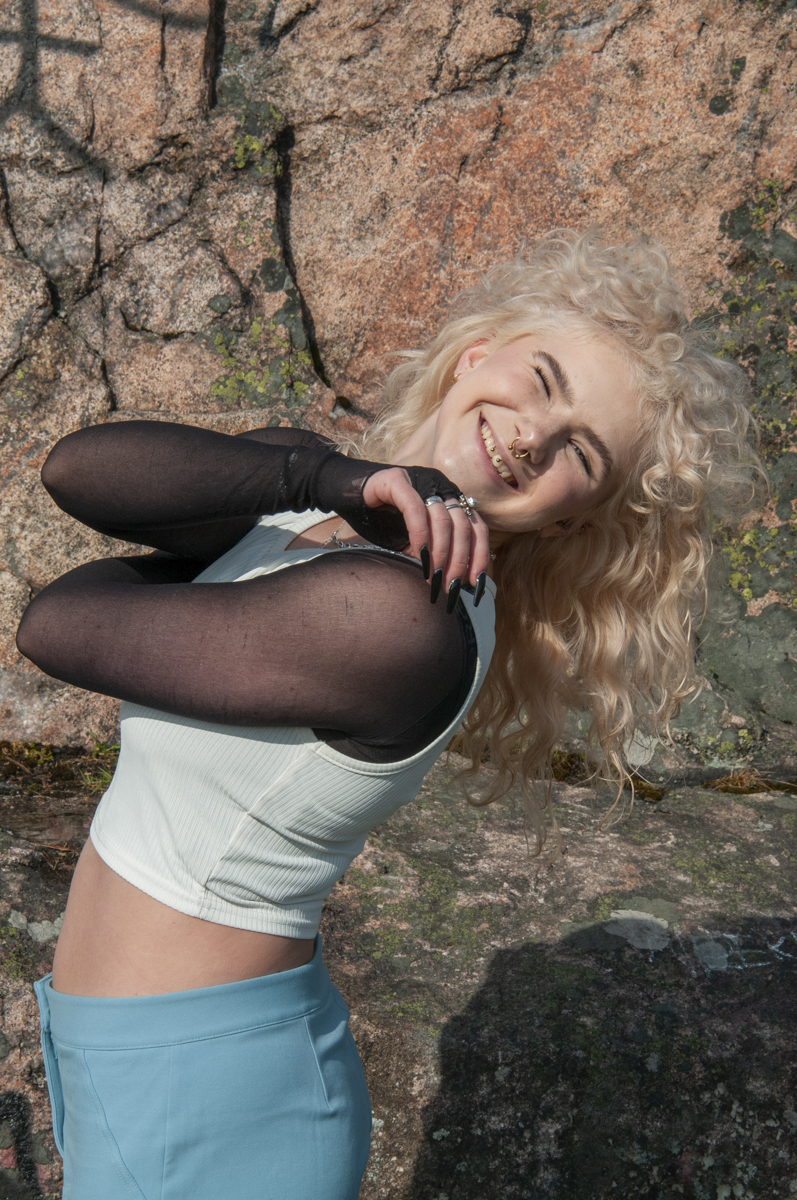 Smiling woman with curly blonde hair wearing a white sleeveless top over a sheer black long-sleeve shirt and blue pants, posing against a rocky background with moss.