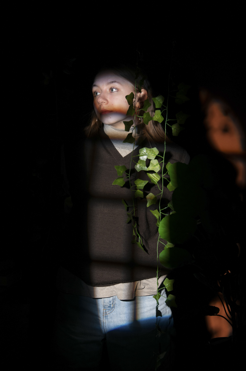 Young woman with light skin and blonde hair wearing a dark sweater and light jeans, standing partially illuminated by a window's shadow grid with green ivy leaves in front of her and a faint blurred reflection to the right.