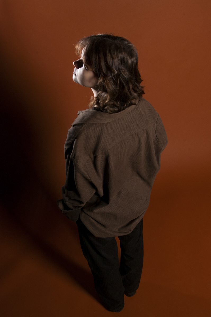 Person with medium-length wavy brown hair and fair skin standing against a solid brown background, wearing a loose brown corduroy shirt and black pants, looking upward and slightly to the side with hands in pockets.