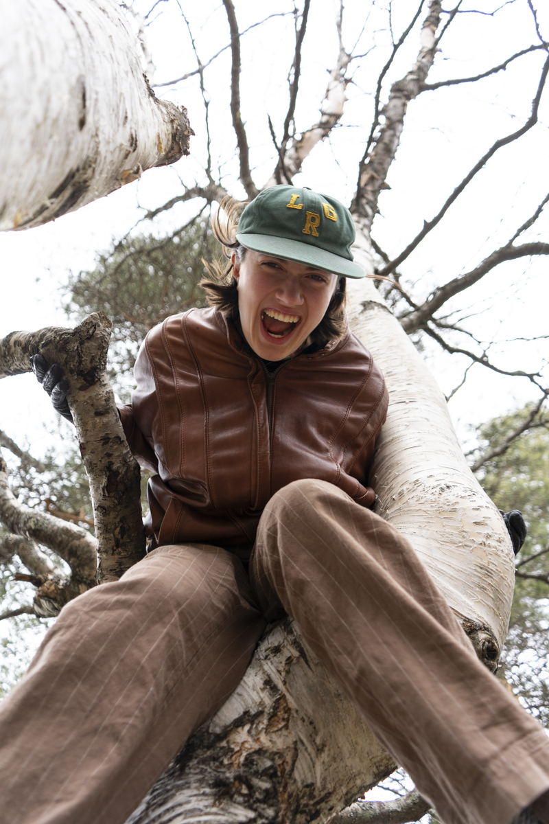 Person wearing a green cap and brown jacket sitting in a tree, smiling widely and holding onto branches with bare limbs in the background.