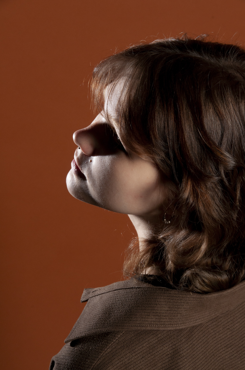 Side profile of a woman with shoulder-length wavy brown hair against an orange background, wearing a brown textured garment and a small hoop earring.
