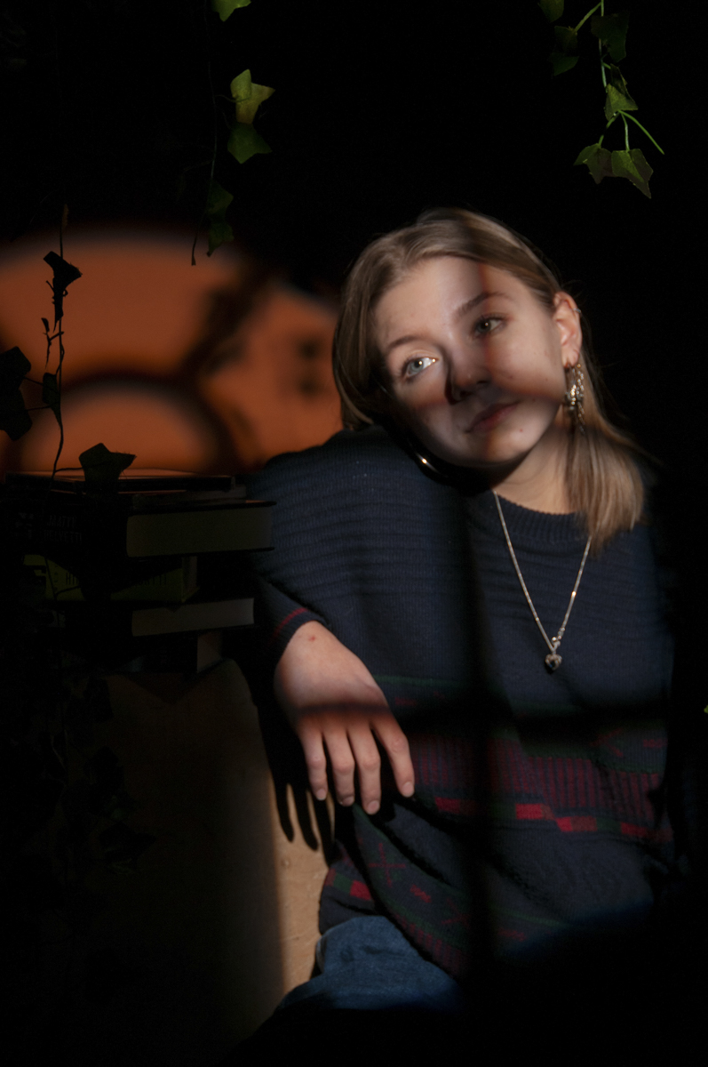 Young woman with light brown hair wearing a dark sweater with red and green patterns, resting her arm and leaning slightly, with shadows cast across her face and a dark blurred background with some greenery and indistinct shapes.