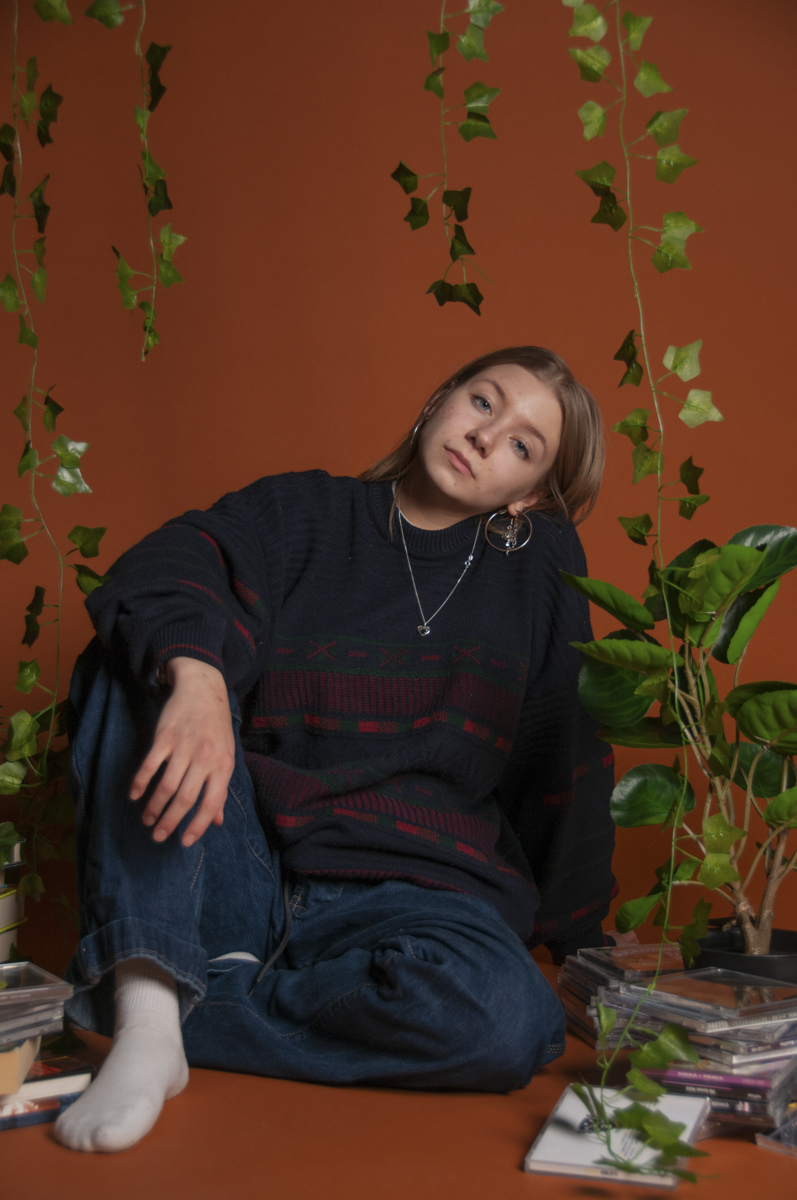 Young woman with light brown hair sitting on a rust-colored floor against a rust-colored background, wearing a dark sweater and blue jeans with white socks, surrounded by green hanging and potted plants and stacks of CDs.