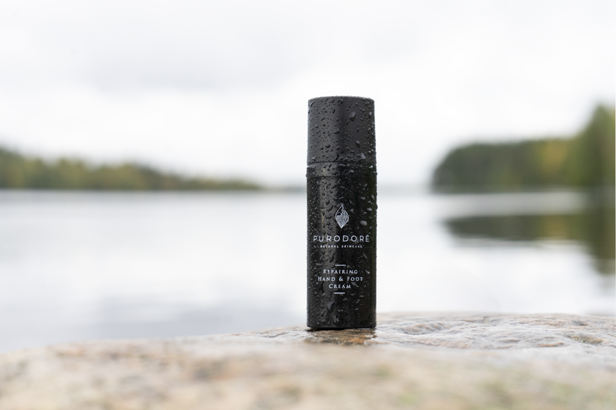 Black cylindrical bottle of Purodoré Repairing Hand & Foot Cream with droplets on its surface, placed on a rock by a calm lake with blurred trees and sky in the background.