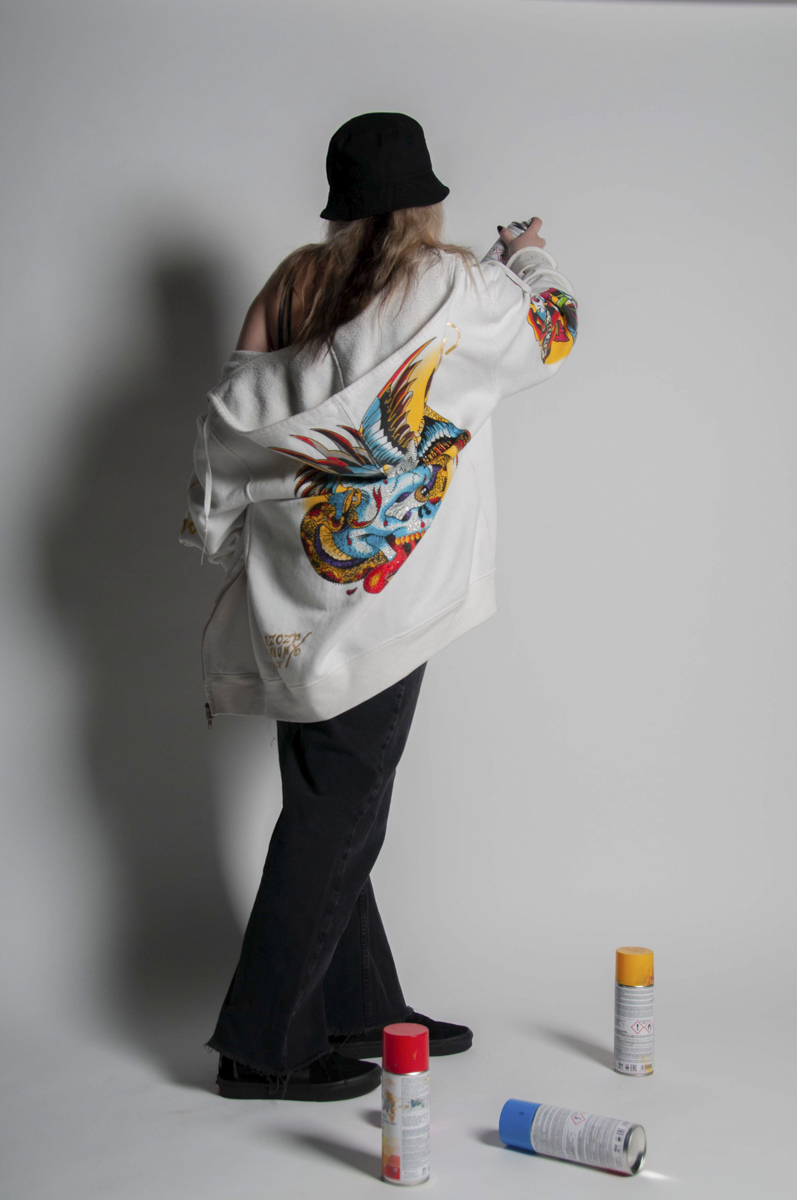 Person wearing a black bucket hat, black pants, and an oversized white jacket with a colorful bird and snake design on the back, holding a can of spray paint while standing on a gray background with three spray paint cans (red, blue, and orange) on the floor.