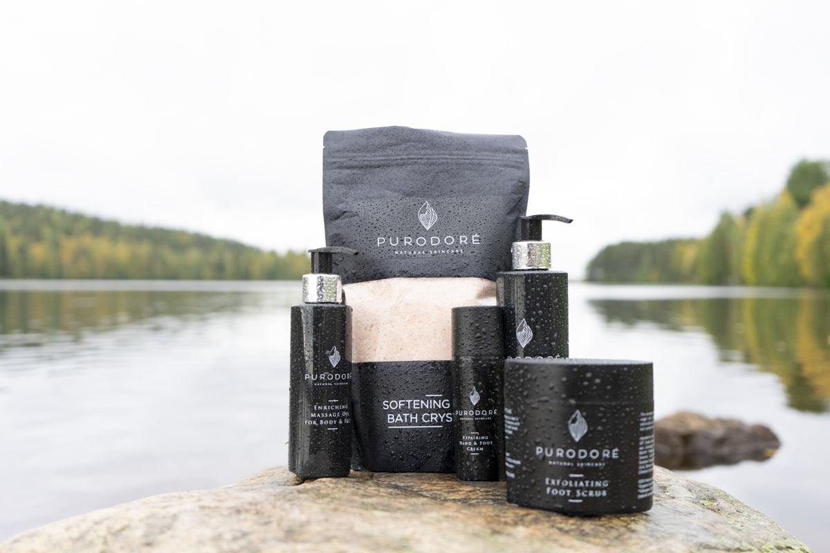 Five PURODORÉ natural skincare products with water droplets on black packaging, including softening bath crystals, enriching massage oil, repairing hand and foot cream, and exfoliating foot scrub, arranged on a rock by a calm lake with forest in background.