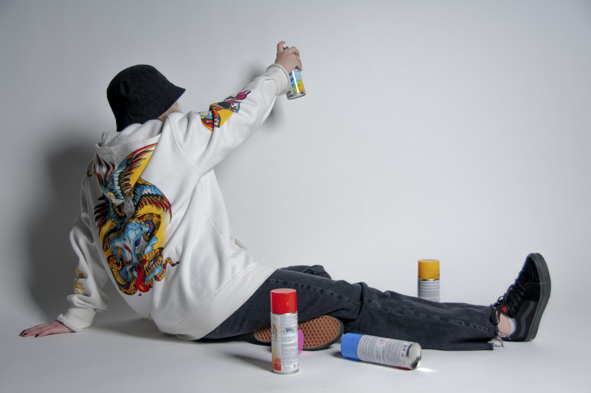 Person sitting on the floor against a white background, wearing a white hoodie with a colorful graphic on the back, black pants, black bucket hat, and black sneakers, holding a spray paint can in one hand with several other spray cans on the floor around them.