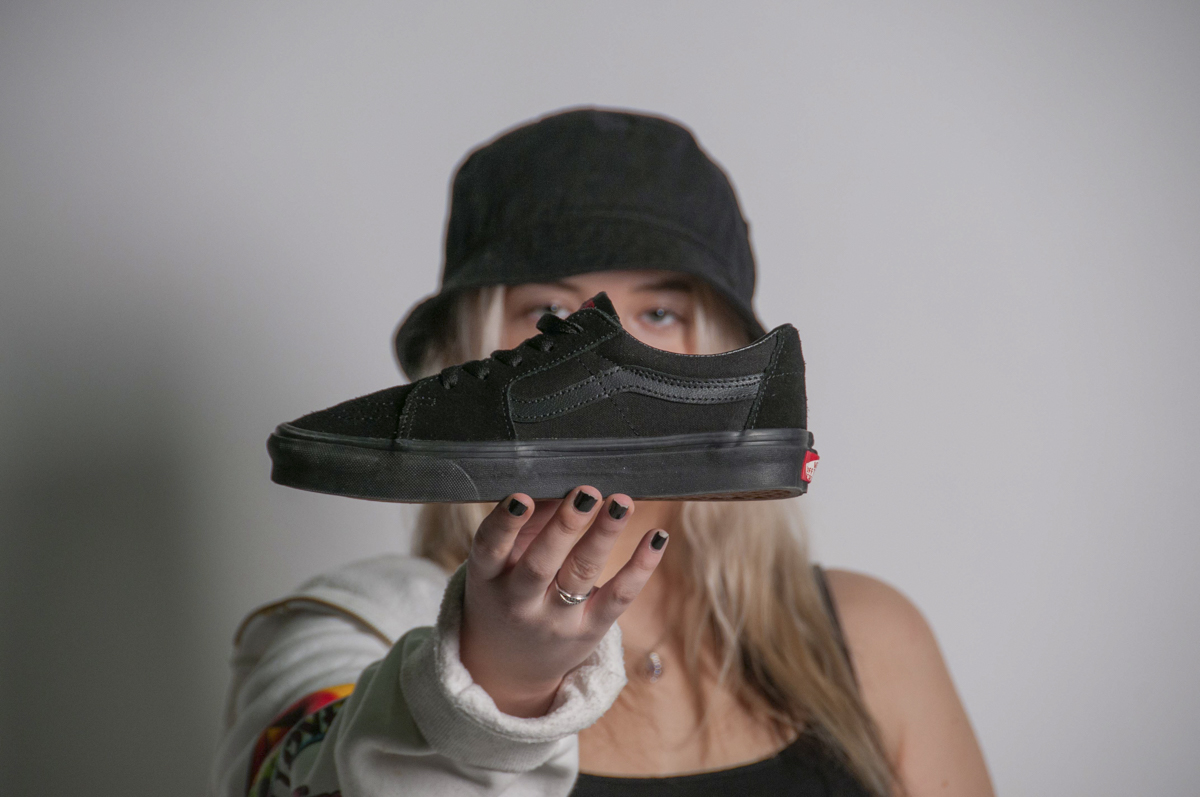 Person wearing a black bucket hat and light-colored hoodie holds a black Vans sneaker toward the camera with black painted nails.