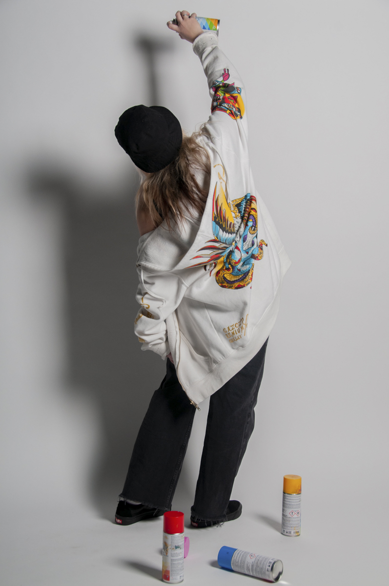 Person wearing a white hoodie with a colorful dragon design on the back, a black beanie, and black pants, spray painting against a gray wall with three spray paint cans on the floor.