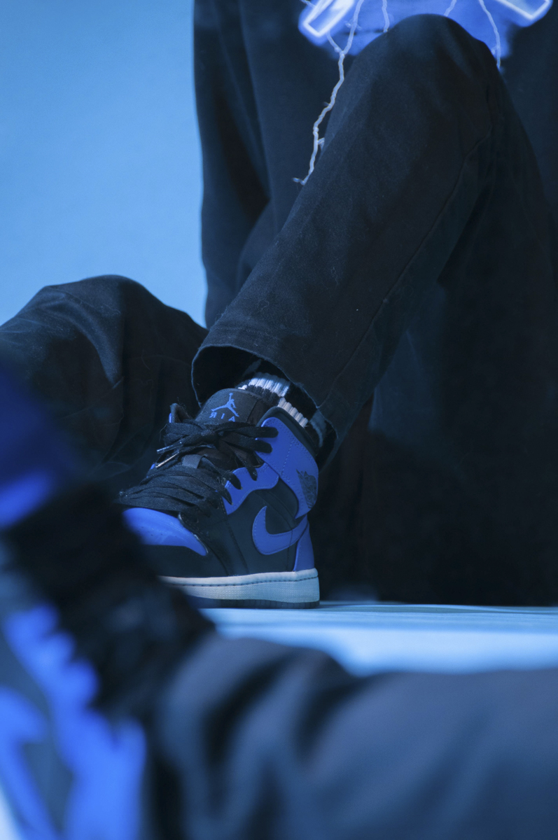 Person wearing black pants and black and blue Nike Air Jordan sneakers with black laces, sitting with one leg bent and resting on the floor under blue lighting.