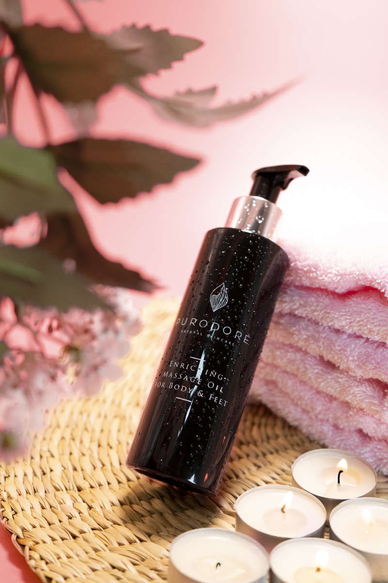 Black bottle of Purodore enriching massage oil for body and feet with water droplets on it, placed on a woven mat beside lit tealight candles and pink folded towels, with blurred green leaves in background.