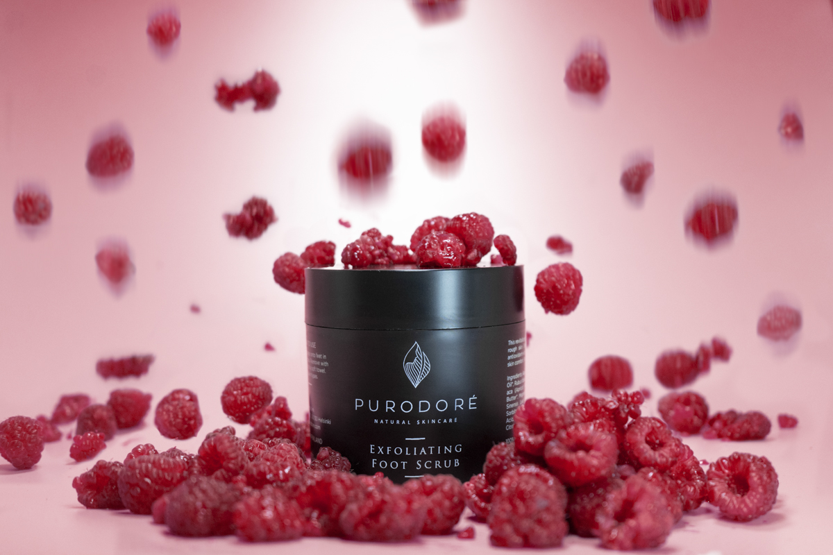 Black container of Purodoré natural skincare exfoliating foot scrub surrounded by and topped with fresh raspberries against a pink background.